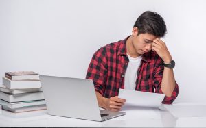 A male student sitting at a desk with a laptop and a stack of books
